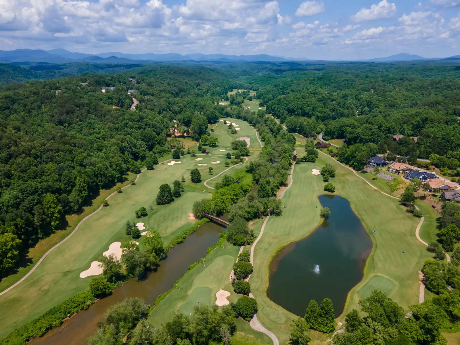 Aerial view of golf fairways with sand bunkers and a fountain pond winding through dense hardwood forest, with homes along the tree line and the blue ridgelines of the North Georgia mountains on the horizon