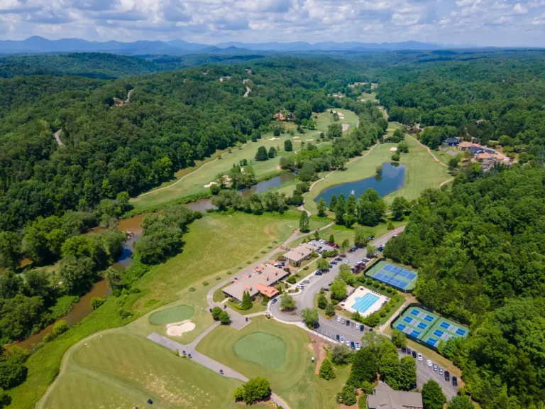Aerial drone view of Achasta Golf Community in Dahlonega, GA showing the clubhouse with terracotta roof, practice putting greens, community pool, tennis and pickleball courts, golf fairways with two ponds, the Chestatee River, homes along the fairway, and forested Blue Ridge foothills on the horizon.