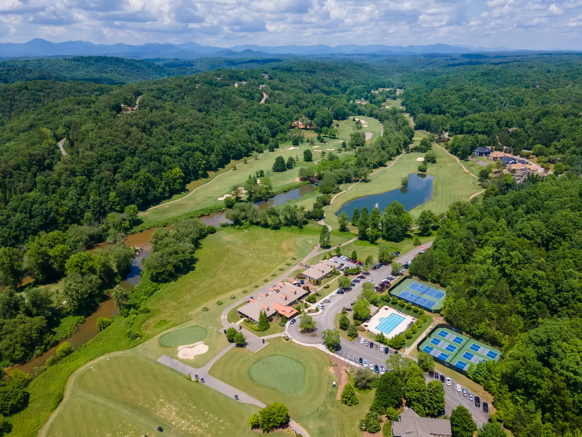 Aerial drone view of Achasta Golf Community in Dahlonega, GA showing the clubhouse with terracotta roof, practice putting greens, community pool, tennis and pickleball courts, golf fairways with two ponds, the Chestatee River, homes along the fairway, and forested Blue Ridge foothills on the horizon.